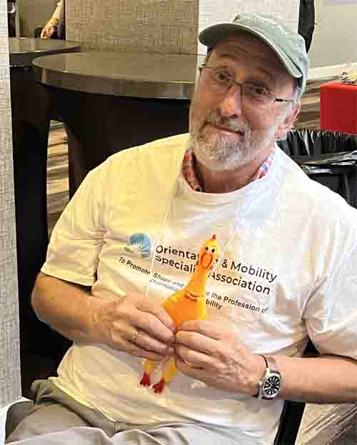 Lukas sits and smiles, wearing his O&M Specialist Association shirt and holding a yellor rubber chicken.